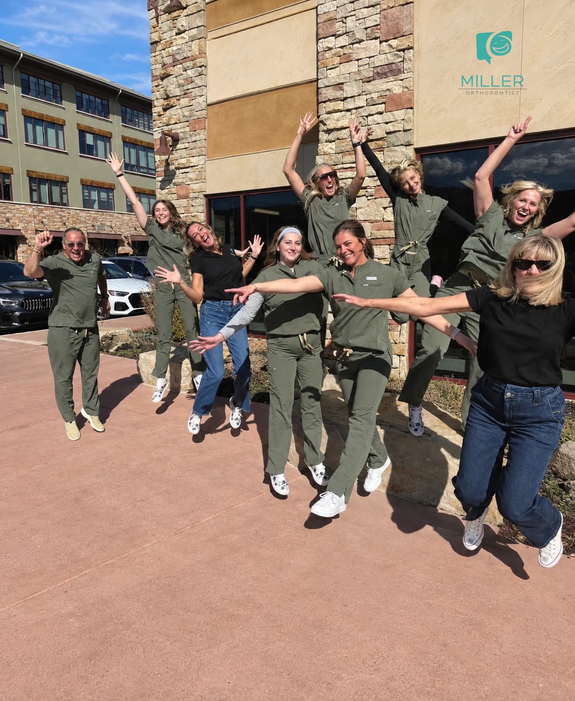 The Miller Orthodontics team in matching green scrubs jumping for joy outside the office, with the Miller Orthodontics logo on the wall